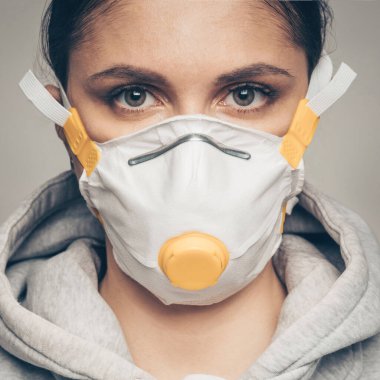 Stüdyo portresi, yüz maskesi takan, kameraya bakan, gri arkaplana yakın genç bir kadın. Coronavirus salgını, toz alerjisi, virüs konseptine karşı koruma