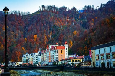 Nehir kıyısındaki güzel, modern binalar Rosa Khutor 'un kayak merkezindeki parlak sarı ağaçlarla kaplı dağların arka planına karşı.,