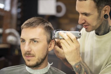 Berber, berber dükkanında erkek müşteriye tıraş olurken elektrikli tıraş makinesi kullanıyor.