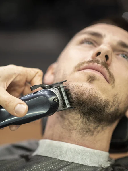 Barber using electric razor while trimming a beard of male customer at barber shop