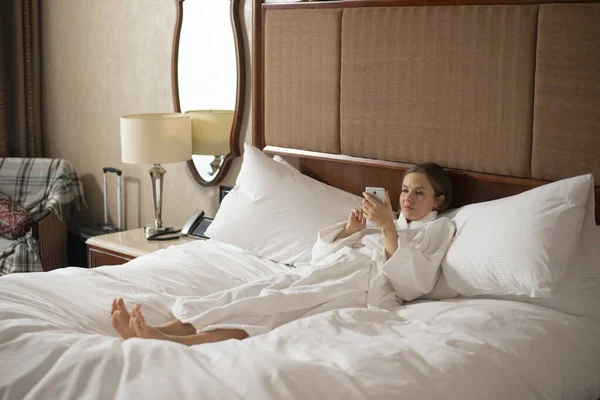 Donna Accappatoio Bianco Sdraiata Sul Letto Con Smartphone — Foto Stock