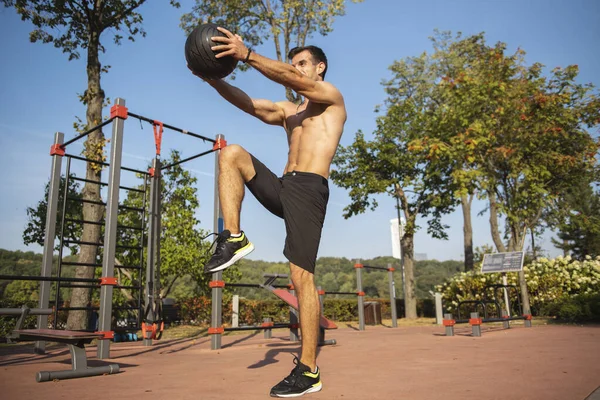Fit guy doing exercises using a ball outdoors. Young athletic man training in city park