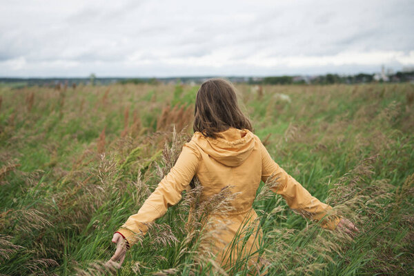 Young woman in yellow jacket with opened arms standing in field against cloudy sky looking at horizon