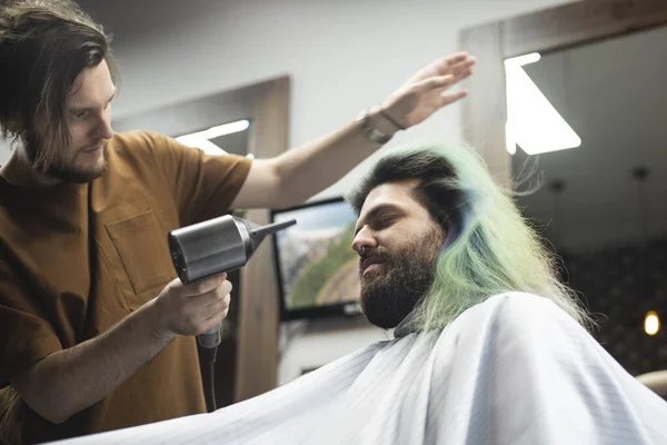 Male customer sitting in barber chair with closed eyes enjoying the providing of hairstyle. Barber using a hair dryer for hairstyle making