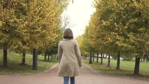Женщина в пальто гуляет по осеннему парку вдоль деревьев — стоковое видео