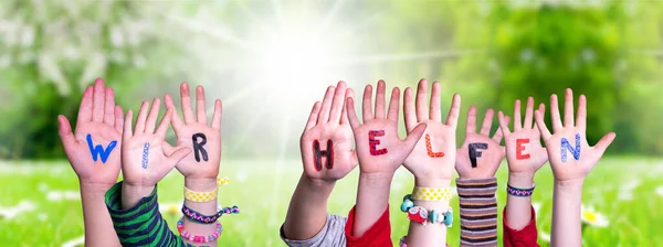 Las manos de los niños sosteniendo la palabra Wir Helfen significa que ayudamos, prado de hierba — Foto de Stock