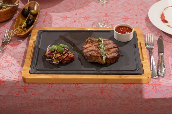 Sliced prime ribeye steak on black stone plate. Medium degree of steak doneness. With rosemary and peppers. Fork and knife. — Stock Photo, Image