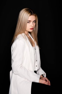 Genç, güzel bir kız, siyah arka planda siyah pantolon ve beyaz tişörtle bir sandalyeye oturmuş poz veriyor. Kameraya bak..