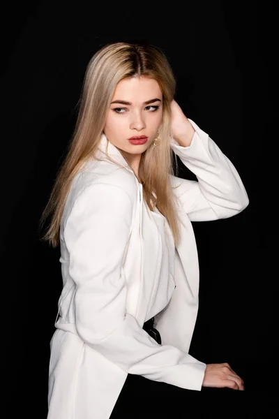 Young beautiful girl posing sitting on a chair in a white t-shirt and black trousers on a black background. Looks at the camera. — Stock Photo, Image