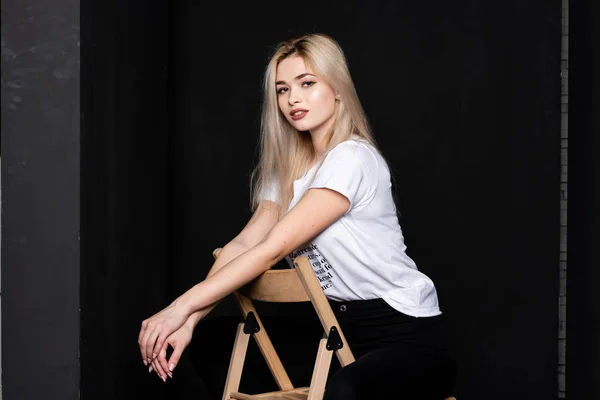 Young beautiful girl posing sitting on a chair in a white t-shirt and black trousers on a black background. Looks at the camera. — Stock Photo, Image