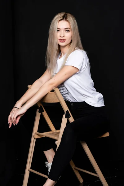 Ung vacker flicka poserar på en stol i en vit t-shirt och svarta byxor på en svart bakgrund. Titta på kameran.. — Stockfoto