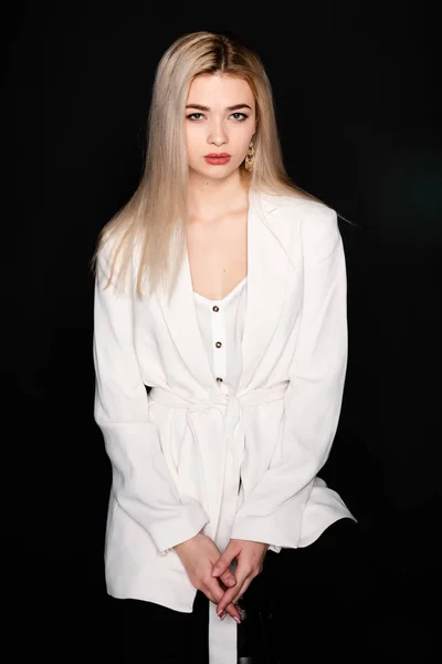 Joven chica hermosa posando sentado en una silla en una camiseta blanca y pantalones negros sobre un fondo negro. Mira la cámara. . — Foto de Stock