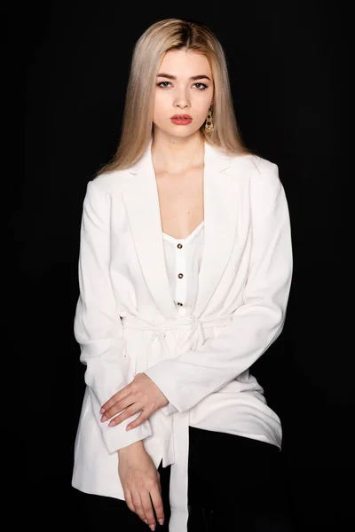 Young beautiful girl posing sitting on a chair in a white t-shirt and black trousers on a black background. Looks at the camera. — Stock Photo, Image