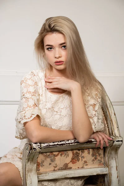 Retrato de uma jovem linda loira com cabelo comprido. Uma menina bonita em um vestido de renda branco senta-se em uma cadeira vintage contra uma parede branca com sombras geométricas . — Fotografia de Stock