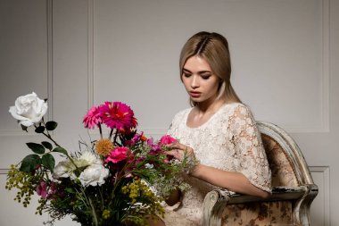 Uzun saçlı güzel bir sarışının portresi. Beyaz dantel elbiseli güzel bir kız, eski bir sandalyede oturuyor. Geometrik gölgeli beyaz bir duvara yaslanmış..