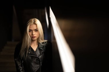 Siyah deri ceketli güzel sarışın bir kızın portresi. Merdiven basamaklarında ışık altında oturuyor. Metin için boş alan