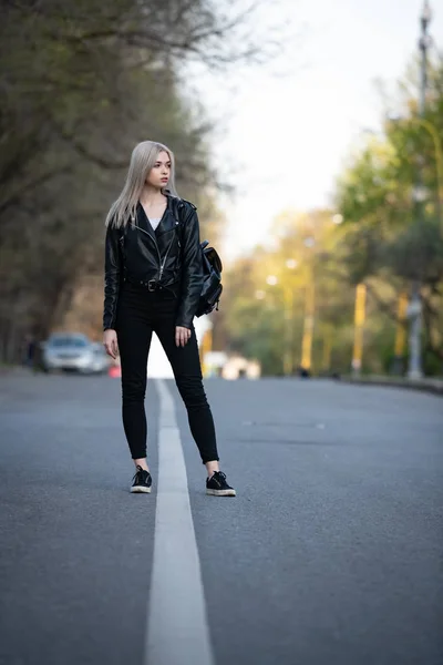 Una giovane bella ragazza bionda con una giacca di pelle nera cammina lungo una strada di città vuota, l'angolo inferiore della sparatoria — Foto Stock