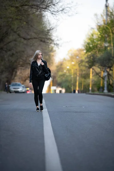 Молодая красивая блондинка в черной кожаной куртке идет по пустой городской дороге, под нижним углом стрельбы — стоковое фото