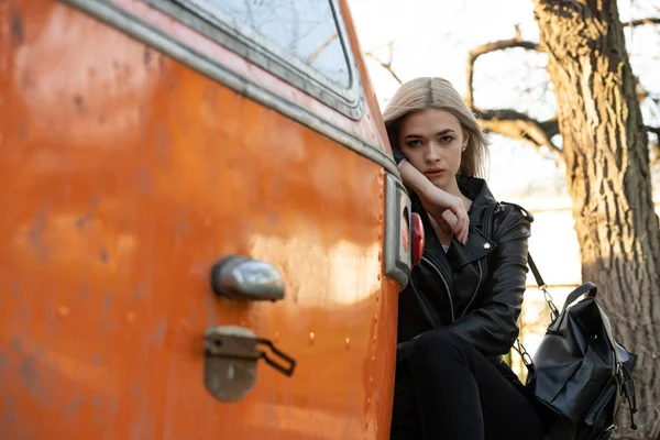 一位穿着黑色皮夹克的金发姑娘站在一辆橙色面包车旁边 — 图库照片