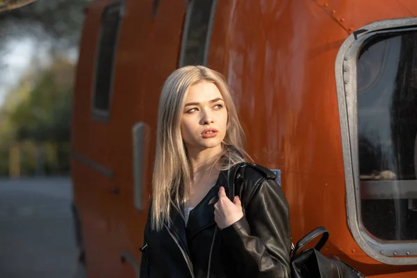 Een jong mooi blond meisje in een zwart leren jasje staat naast een oranje busje — Stockfoto