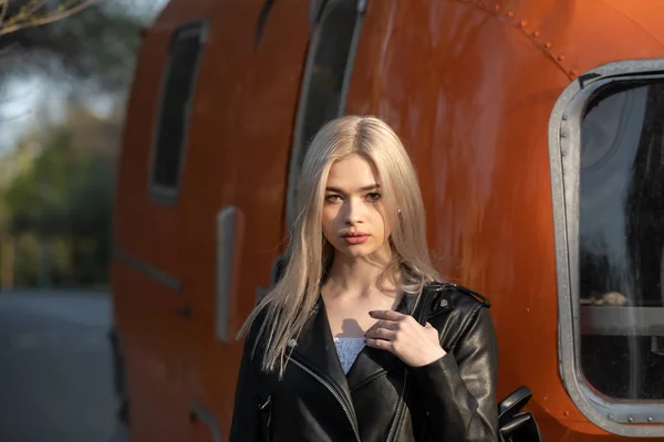 Uma jovem linda garota loira em uma jaqueta de couro preto fica ao lado de uma van laranja — Fotografia de Stock