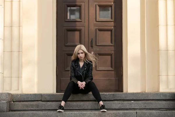 A beautiful young blonde girl stands in a black leather jacket on the background of a building — Stock Photo, Image