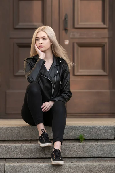 Piękna młoda blondynka stoi w czarnej skórzanej kurtce na tle budynku — Zdjęcie stockowe