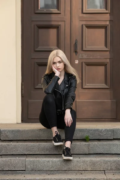 Krásná mladá blondýna dívka stojí v černé kožené bundě na pozadí budovy — Stock fotografie