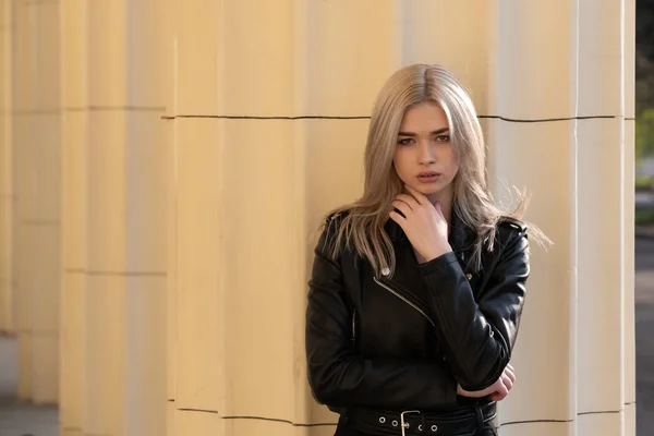 A beautiful young blonde girl stands in a black leather jacket on the background of a building — 스톡 사진