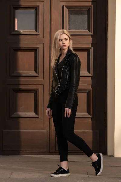 A beautiful young blonde girl stands in a black leather jacket on the background of a building — 스톡 사진