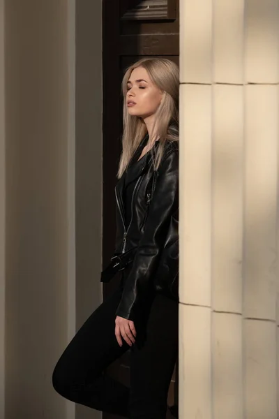 A beautiful young blonde girl stands in a black leather jacket on the background of a building — 스톡 사진