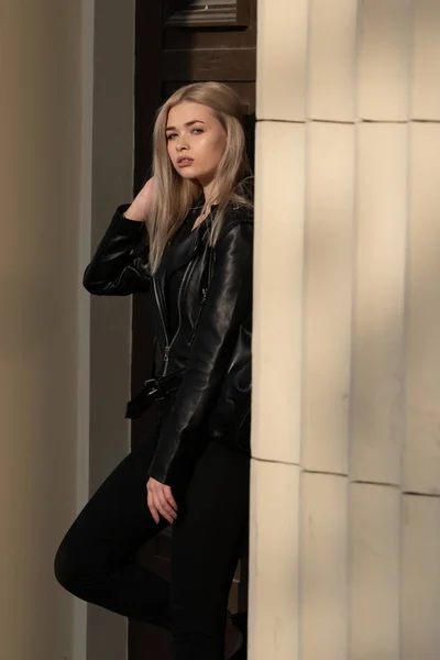 A beautiful young blonde girl stands in a black leather jacket on the background of a building — 스톡 사진