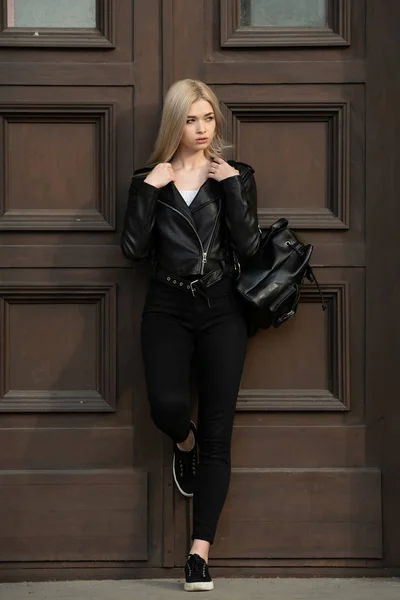 一个漂亮的金发姑娘穿着黑色皮夹克站在一座大楼的后面 — 图库照片