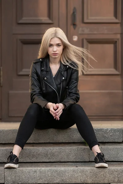 A beautiful young blonde girl stands in a black leather jacket on the background of a building — Stock Photo, Image