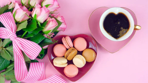 Mothers Day coffee and macarons with roses gift overhead on pink wood table.