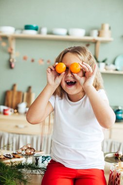 Gözlerinde mandalina olan neşeli küçük bir kızın portresi, Noel, mutfak, rahat bir ev.