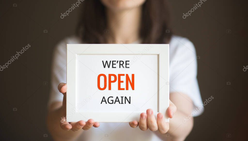 text we are open again. person holds white frame in her hands. woman in white t-shirt on dark background.