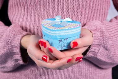 Kalp şeklinde mavi bir hediye kutusu ve arka planda pembe yün bir kazak tutan kız elleri. Parmakları kırmızı ojeli kadın elleri. Noel ya da Sevgililer Günü tatili konsepti