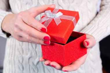 El ele tutuşup kırmızı hediye kutusunu açan ve arka planda yün süveter giyen bir kız. Parmakları kırmızı ojeli kadın elleri. Noel ya da Sevgililer Günü tatili konsepti.