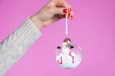 Stüdyoda pembe arka planda karlı bir süs topu tutan kız. Parmakları kırmızı ojeli kadın elleri. Noel ya da Sevgililer Günü bayramı ve minimalizm kavramı