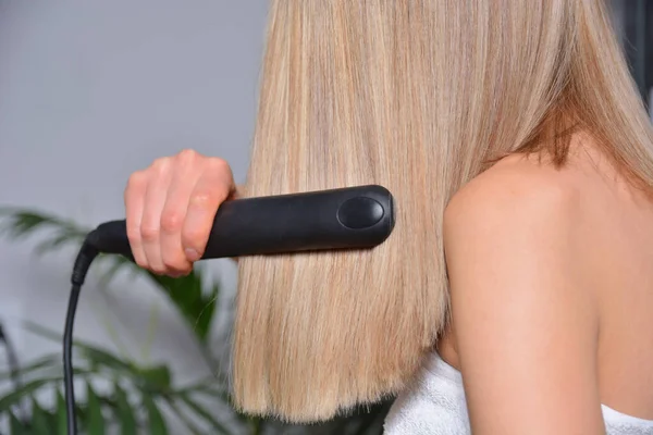 Woman ironing long blonde hair with a flat iron at home. Cares about healthy and clean hair. Girl wears a white towel. Female with bare shoulders. Beauty salon concept