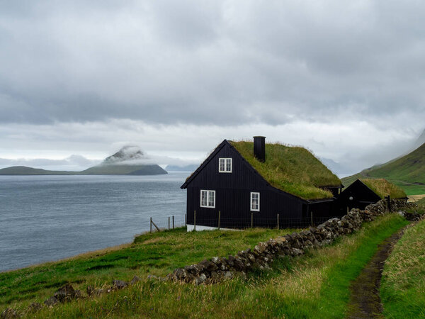 Faroe Islands. House with grass roof on the slope of hill. Hazy landscape. Cloudy weather.
