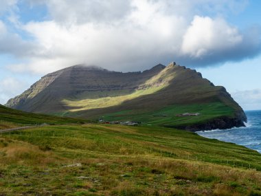 Faroe Adaları, Vidoy, Vidaredii. Uçurumların ve köyün üzerindeki yeşil tepelerin muhteşem manzarası. Mavi gökyüzü biraz bulutlu.