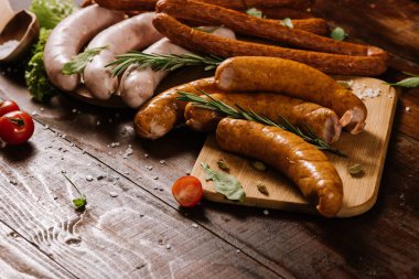 Farklı tütsülenmiş sosisler, sebzeler ve ahşap arka planda malzemeler.