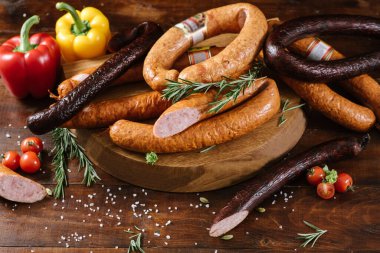 Farklı tütsülenmiş sosisler, sebzeler ve ahşap arka planda malzemeler.