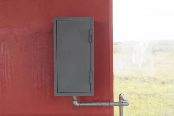 Szare żelazne pudełko wisi na czerwonej ścianie, obok okna. Skrzynka strażacka lub gazowa, przewód wodny lub gazowy jest do niej podłączony. — Zdjęcie stockowe