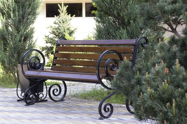 Dřevěná hnědá lavička se železnými kulatými ozdobnými rukojetěmi v parku. Zelené smrkové větve, zábavní park pro lidi, zelené rostliny. Hotel má rekreační areál v parku v sanatoriu. — Stock fotografie