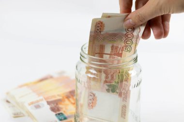 El, cam bir kavanozda 5000 ruble 'lik bir destenin içine Rus rublesi koyuyor. Bir tomar parayla izole edilmiş beyaz bir arka plan. Tasarruf ve finans birikimi kavramı.
