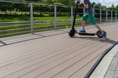 Genç bir çocuk sokakta elektrikli scooter kullanıyor. Şehir parkı, yaz güneşli bir gün, nehir kenarındaki ahşap gezinti alanı. Modern şehir taşımacılığı. Çocuk ayağını scooter 'a basıyor..