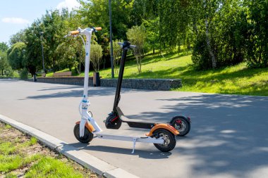 İki elektrikli scooter, siyah ve beyaz, caddedeki tramvayda duruyor. Güneşli bir yaz günü. Modern şehir taşımacılığı. Ağaçlı şehir parkı.